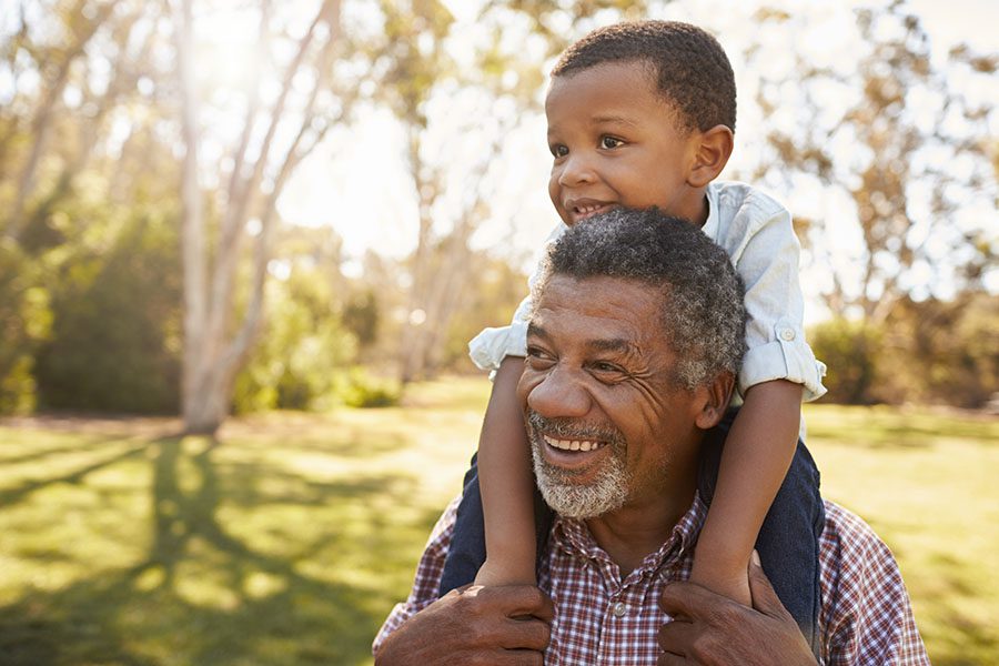 About Our Agency - Closeup Portrait of a Cheerful Grandfather Giving His Grandson a Piggyback Ride Outside in the Backyard on Warm Sunny Day