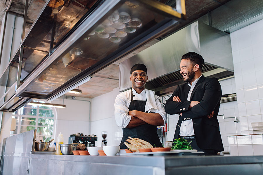 Specialized Business Insurance - View of a Smiling Restaurant Manager and Cook Standing Behind the Counter in the Kitchen of a Restaurant